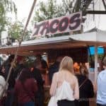 Eine Menschenmenge steht vor einem Taco-Stand im Freien mit einem großen 'Tacos'-Schild.