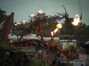 Eine Drachen-Skulptur aus Metall speit Flammen in einem Garten mit Bäumen und Zelten im Hintergrund.