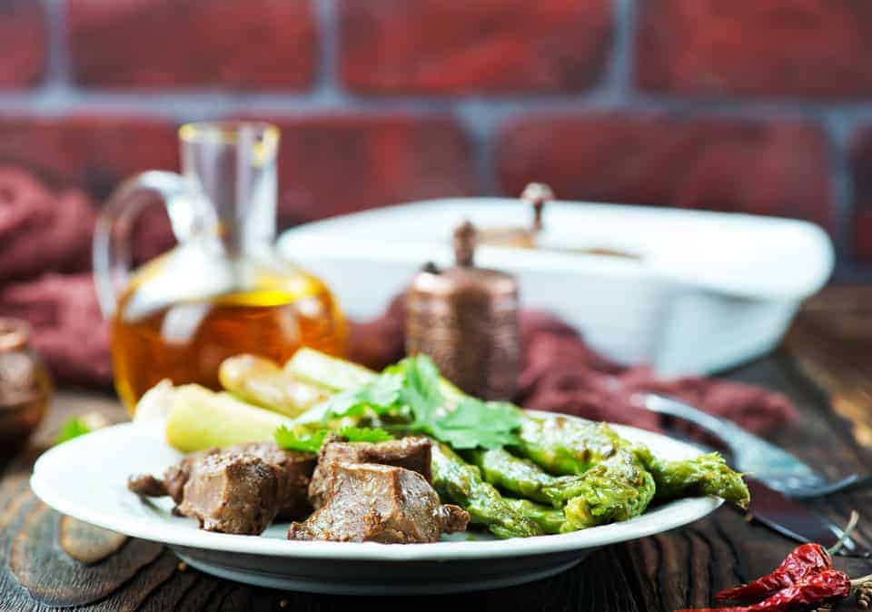 Ein Teller mit Fleisch, Spargel und Kräutern, daneben eine Karaffe Öl auf einem Holztisch.