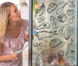 Frau betrachtet Zeichnungen von Lebensmitteln auf einer Glasscheibe, darunter Bananen, Fisch und Käse.