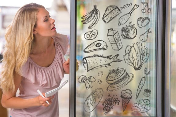 Frau betrachtet Zeichnungen von Lebensmitteln auf einer Glasscheibe, darunter Bananen, Fisch und Käse.