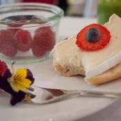 Ein belegtes Brötchen mit Käse, Erdbeere und Heidelbeere neben Himbeeren und essbaren Blüten auf einem Teller.