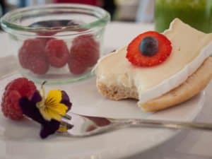 Ein belegtes Brötchen mit Käse, Erdbeere und Heidelbeere neben Himbeeren und essbaren Blüten auf einem Teller.