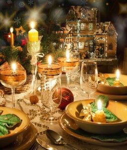 Festlich gedeckter Tisch mit Kerzen, Weihnachtsschmuck und Lebkuchenhaus im Hintergrund.