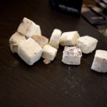 Weiße Marshmallow-Würfel liegen auf einer dunklen Schieferplatte neben einem Glas mit Holzstäbchen.