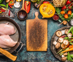 Rohes Hähnchen, Gemüse und Gewürze um ein Holzbrett auf einem rustikalen Tisch.