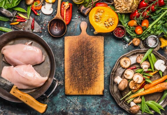Rohes Hähnchen, Gemüse und Gewürze um ein Holzbrett auf einem rustikalen Tisch.