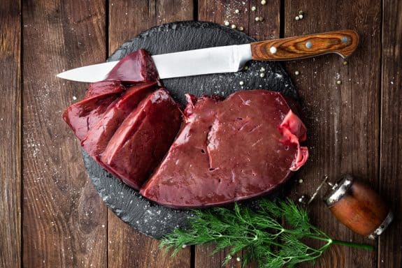 Rohe Leber auf Schieferplatte mit Messer, Pfefferkörnern und Dill auf Holztisch.