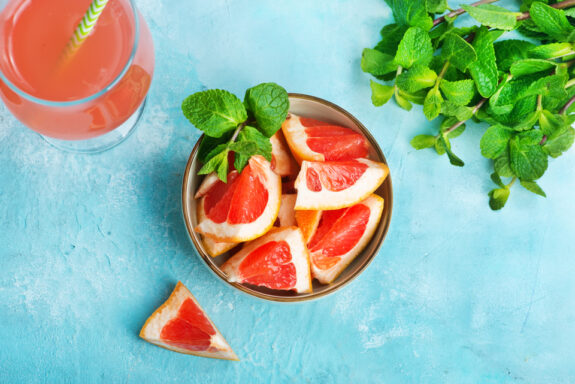 Schüssel mit Grapefruitstücken und Minze, daneben ein Glas Saft und frische Minzblätter auf blauem Hintergrund.