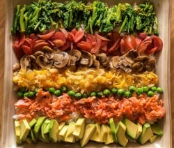 Bunte Gemüsestreifen mit Brokkoli, Paprika, Pilzen, Erbsen, Lachs und Avocado auf einem Tablett arrangiert.