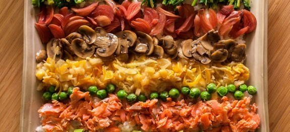 Bunte Gemüsestreifen mit Brokkoli, Paprika, Pilzen, Erbsen, Lachs und Avocado auf einem Tablett arrangiert.