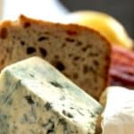 Ein Stück Blauschimmelkäse vor Brot und Wurst auf einem Holzbrett.