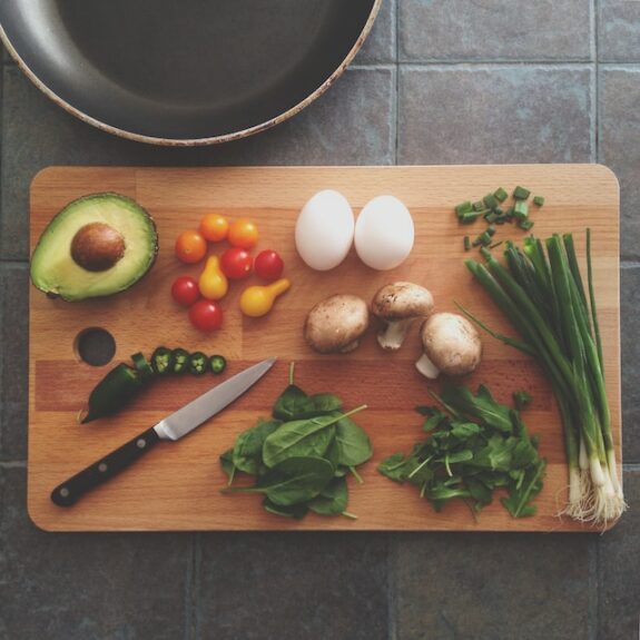 Avocado, Kirschtomaten, Eier, Pilze, Frühlingszwiebeln und Spinat auf Holzbrett mit Messer, Pfanne im Hintergrund.