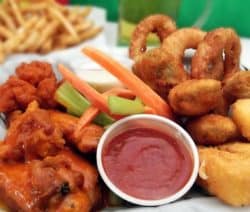 Ein Teller mit Chicken Wings, Gemüse, Zwiebelringen und Dip, im Hintergrund Pommes.