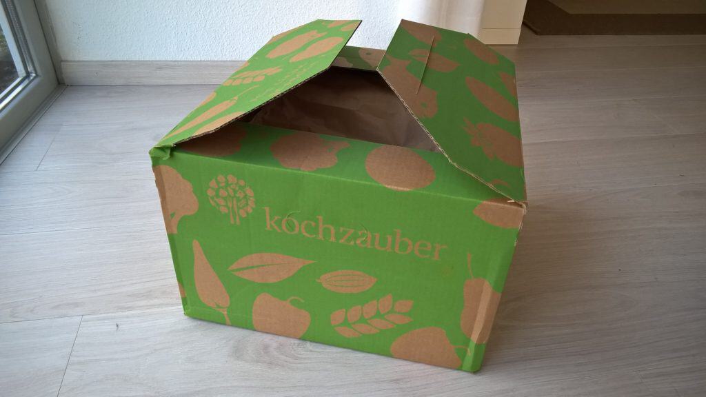 Ein geöffneter, grüner Karton mit braunen Lebensmittelsymbolen steht auf hellem Holzboden.