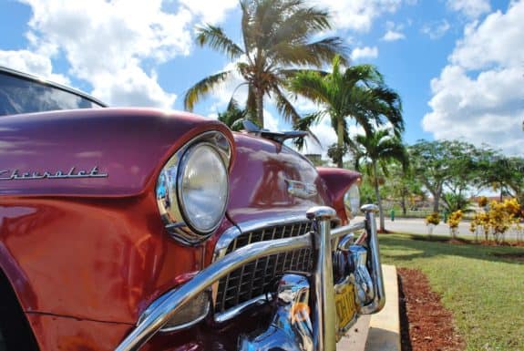 Ein roter Oldtimer vor Palmen und blauem Himmel mit weißen Wolken im Hintergrund.