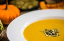 Eine Schüssel Kürbissuppe mit Kürbiskernen, daneben ein grüner und ein orangefarbener Kürbis.