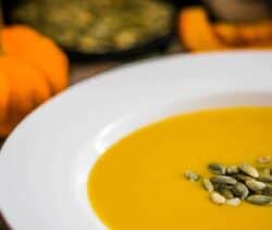 Eine Schüssel Kürbissuppe mit Kürbiskernen, daneben ein grüner und ein orangefarbener Kürbis.