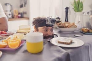 Ein Mann mit Kamera auf dem Kopf schaut lächelnd über einen Frühstückstisch mit Brot, Obst und Käse.