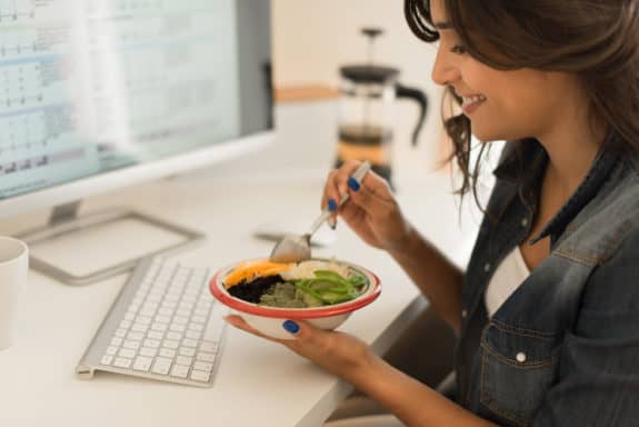 Eine Frau isst Salat am Schreibtisch vor einem Computerbildschirm.