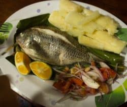 Gekochter Fisch mit Limettenhälften, Yuca-Stücken und Tomaten-Zwiebel-Salat auf einem Teller.