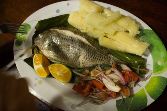 Gekochter Fisch mit Limettenhälften, Yuca-Stücken und Tomaten-Zwiebel-Salat auf einem Teller.