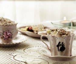 Zwei verzierte Teetassen mit Sahne auf Spitze, dahinter Kekse und brennende Teelichter.