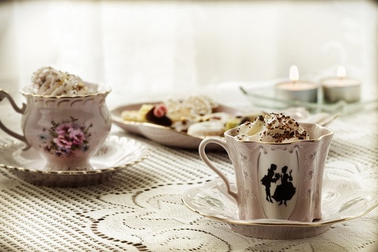 Zwei verzierte Teetassen mit Sahne auf Spitze, dahinter Kekse und brennende Teelichter.