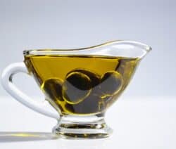 Ein Glasgefäß mit grünem Olivenöl und schwarzen Oliven vor hellem Hintergrund.