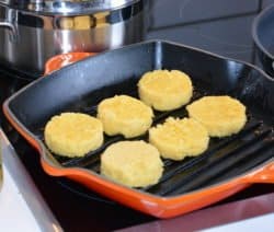 Sechs goldene Polenta-Scheiben braten in einer orangen Grillpfanne auf einem Herd.