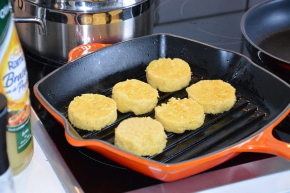 Sechs goldene Polenta-Scheiben braten in einer orangen Grillpfanne auf einem Herd.
