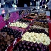 Eine bunte Auswahl an Pralinen auf einem Marktstand mit rotem Tisch und vielen Besuchern im Hintergrund.