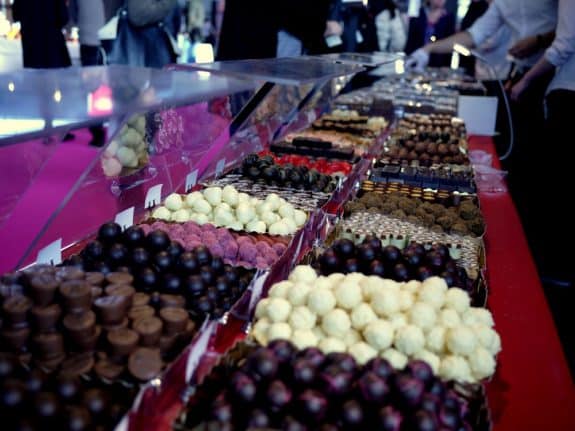 Eine bunte Auswahl an Pralinen auf einem Marktstand mit rotem Tisch und vielen Besuchern im Hintergrund.