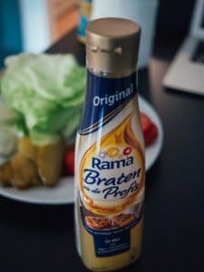 Flasche Rama Bratöl vor unscharfem Salat und Gemüse auf einem Tisch.