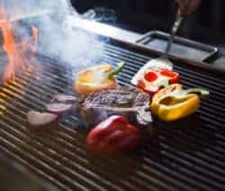 Ein Steak und bunte Paprika grillen neben roten Zwiebeln auf einem heißen Grill mit Flammen und Rauch.