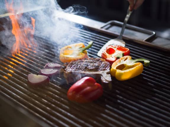 Ein Steak und bunte Paprika grillen neben roten Zwiebeln auf einem heißen Grill mit Flammen und Rauch.