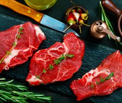 Drei rohe Rindersteaks mit Kräutern auf einer Schieferplatte, daneben ein Messer, Rosmarin und Gewürze.