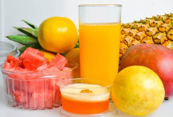 Ein Glas Orangensaft, Ananas, Mango, Melonenstücke und eine Zitrone auf weißem Hintergrund.