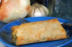 Ein goldbrauner Teigstrudel auf einem blauen Teller, daneben Zwiebel und Knoblauch.