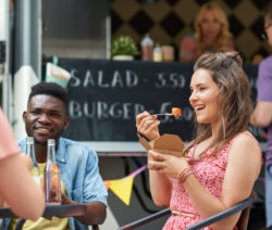 Zwei Personen essen vor einem Foodtruck, eine Frau lacht mit einer Gabel in der Hand.