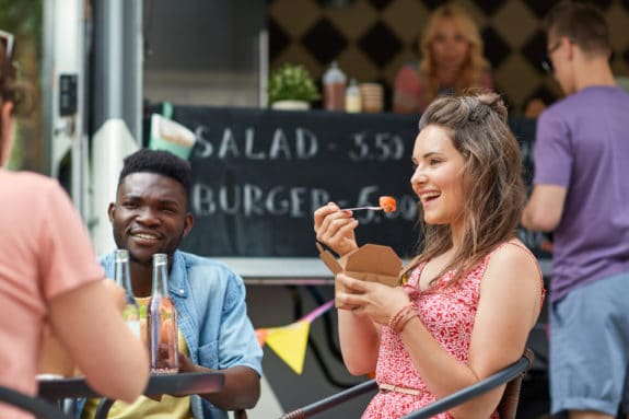 Zwei Personen essen vor einem Foodtruck, eine Frau lacht mit einer Gabel in der Hand.