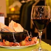 Ein Glas Rotwein neben einem Teller mit Brot, Oliven und Schinken auf einem Tisch.