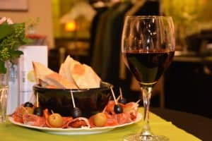 Ein Glas Rotwein neben einem Teller mit Brot, Oliven und Schinken auf einem Tisch.