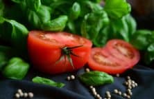 Halbierte Tomate mit Basilikumblättern und weißen Pfefferkörnern auf dunklem Hintergrund.