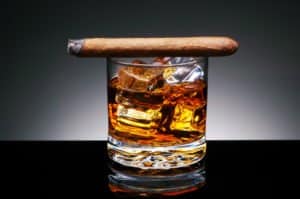 Ein Glas Whisky mit Eiswürfeln, darauf liegt eine angezündete Zigarre.