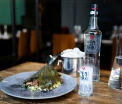 Ein Teller mit gefülltem Weinblatt, daneben eine Yeni Rakı-Flasche und ein Glas auf einem Holztisch.