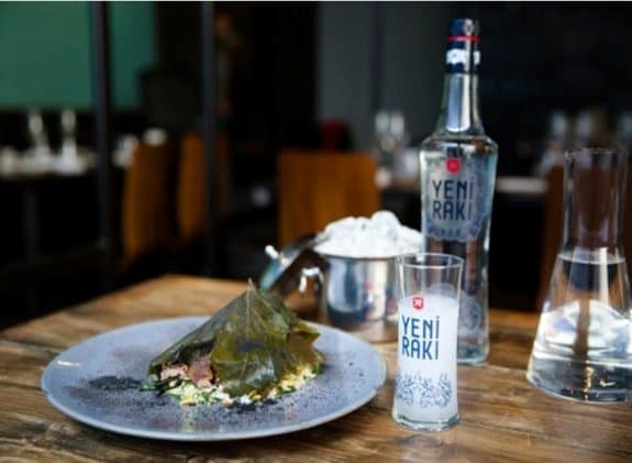 Ein Teller mit gefülltem Weinblatt, daneben eine Yeni Rakı-Flasche und ein Glas auf einem Holztisch.