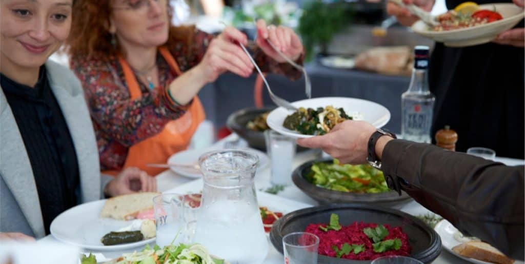 Menschen teilen Essen an einem Tisch mit Salaten, Brot und Wasserkrug.