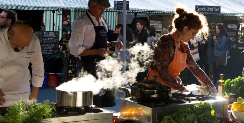 Köche arbeiten an dampfenden Töpfen auf einem belebten Marktstand im Freien.